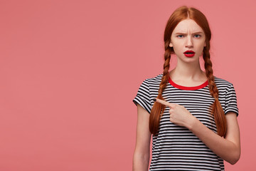 Worried young 20s woman with two red haired braids red lipstick open mouth in despair pointing...