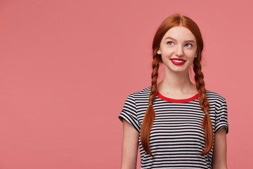 Chrarming pretty beautiful girl with red-haired braids red lips, dressed in stripped t-shirt, smiling dreamingly thoughtfully looks to the upper left corner stands next to the blank copy space