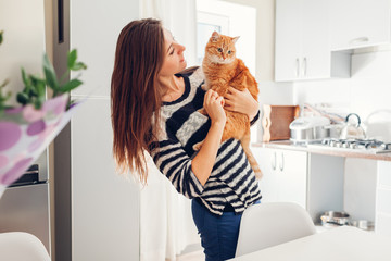 Young woman playing with cat in kitchen at home. Girl holding and hugging ginger cat