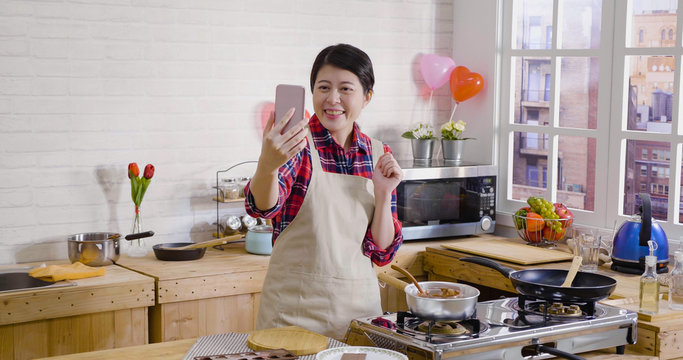 cheerful lady smiling laughing having video phone call on smartphone talking chatting in kitchen while cooking. young asian college girl prepared handmade chocolate for lover. melted choco on stove.