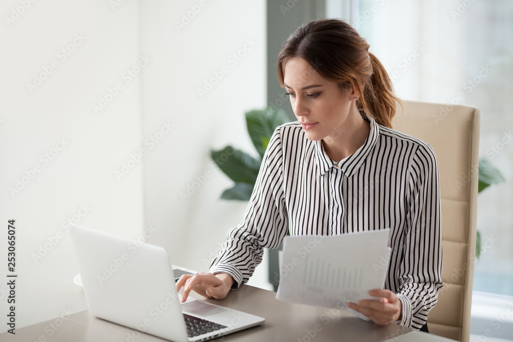 Wall mural serious focused businesswoman typing on laptop holding papers preparing report