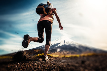 Young woman, trail running athlete runs on the trail with loose ground and volcano on the...