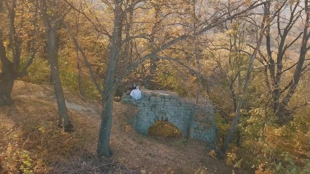 Wedding couple. Lovely groom and bride sitting on on a remains of a destroyed building in the forest.  Happy family. Man and woman in love. Wedding day. Slow motion. Arial view. Shot from drone