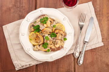 A closeup of scallopine di vitello, veal scallopini, with a mushroom sauce, lemons, and parsley, shot from the top on a dark rustic wooden table with red wine and a place for text