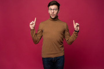 Portrait of a happy young man pointing fingers up at copy space isolated over pink background.