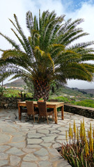 Fototapeta na wymiar Wooden table and chairs beneath a mature tropical palm tree overlooking rustic nature and fincas. Picnic or eating area with a view in a high up village mountains of El Hierro, Canary Islands, Spain.