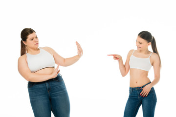 smiling fit woman pointing with finger at overweight sad woman doing stop gesture isolated on white