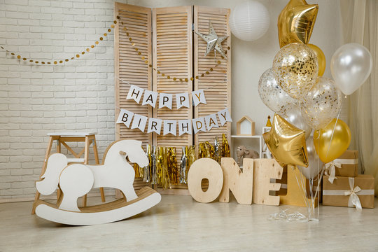 Birthday gold and silver decorations with gifts, toys, garlands and figure for little baby party on a white bricks background.