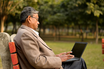 Side view of senior Indian man wearing a tweed coat working on a laptop on a park bench sitting cross legged in New Delhi, India