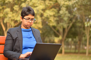 Confident Indian professional business woman in business attire working on a laptop on a park bench