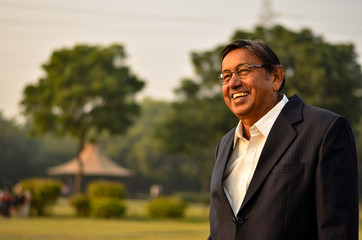 Happy looking senior Indian man wearing a suit standing and smiling, laughing in a park in Delhi, India