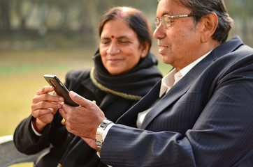 Portrait of a senior Indian couple sitting in park working on their smart phone in Delhi