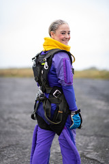 Pretty woman happy, dressed in jumpsuit and parachute harness on airport runway ready for skydive...
