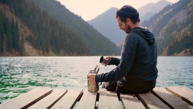 Hipster traveler man sitting on wooden bridge of lake green mountains in connection with nature pouring drinking and enjoying taste of hot tea or thermos beverage Feel calm state of mind in relax