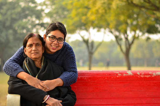 Young Indian woman hugging and smiling with her mother who is sitting on a red bench in a park in New Delhi, India. Concept Mother's day