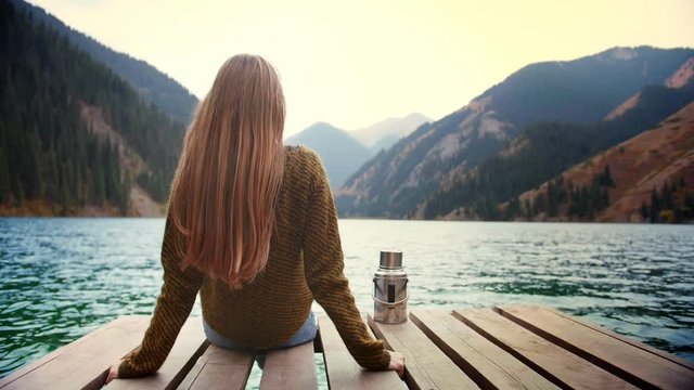 Long hair blond female traveler is doing mindful meditation sitting on bridge of lake green mountains in connection with nature Feel calm state of mind in relax moment of internal mental equilibrium