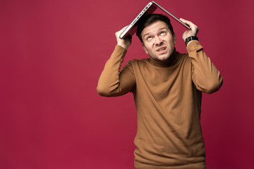 Portrait of unhappy stressed young bearded man holding laptop above head while stsanding isolated over pink background.