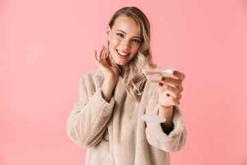 Emotional young pretty woman posing isolated over pink wall background holding glass with alcohol drink.