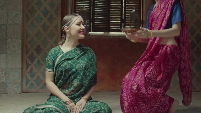 Hospitable female in hindu dress sari with bindi and bracelets serving food to woman guest with tika decoration, seated on floor. Woman offering to try traditional indian meal, Navratan mix to friend.