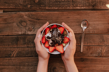 hands holding strawberries with yogurt chocolate pumpkin seeds chia sunflower seeds and apple in a white bowl on a wooden table