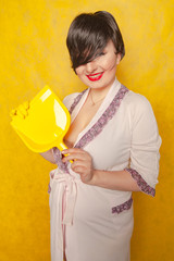 plump woman with short black hair stands in a pink Bathrobe with a rubber cleaning glove on her hand and a dustpan on a yellow Studio background
