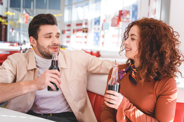 beautiful couple smiling and holding glass bottles with soda at cafe