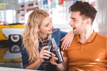 beautiful couple smiling, hugging and cheering with glass bottles at cafe