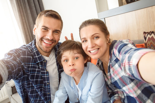Family at home sitting on couch in living room together taking selfie pictures parents making horns to son cheerful close-up