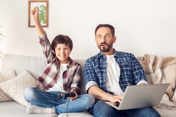 Father and little son at home sitting on sofa dad using laptop looking at tv shocked while boy holding controller hand up cheerful