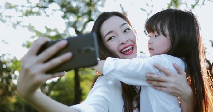 Portrait of mother and her daughter girl using smartphone for selfie at park. Laughing mom and daughter posing for camera in sunset time on summer day