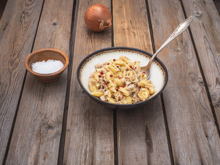 Pasta with minced turkey in a deep bowl and a tablespoon on a wooden plank table, in the background unpeeled onion