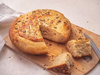 A slice of homemade homemade bread with a sprinkling of sunflower seeds and sesame and reflected loaves of bread are on the kitchen unpainted board.
