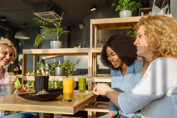 Three happy women multi ethnic friends sitting and chatting in restaurant during lunch....