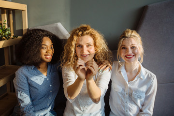 Three happy women holding hands in the form of heart multi ethnic friends sitting beside and chatting in restaurant during lunch. Multi-racial, difference cultures friendship concept.
