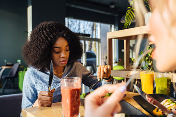 African blavk woman eating lunch or dinner at small cozy cafe. Attractive girl having fun and talking about interesting things with her unrecognizable friends. Multi-racial friendship concept.