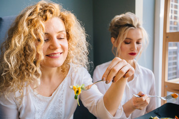 Blond curly woman eating fresh salad at restaurant. Healthy american girl with friend eating salad together looking away. Smiling young woman holding a forkful of salad. Health and diet concept.