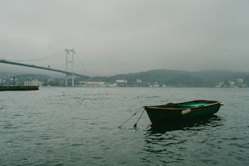 Empty wooden boat near Bosphorus Bridge at Ortakoy district in Istanbul, Turkey. Dramatic mood. The 15 July Martyrs Bridge in a fog