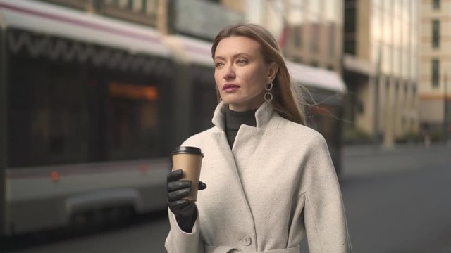Slow motion attractive woman long hair dressed in white coat and grey sweater holding a cup of coffee waiting for the taxi near the roadway in the wind tram on the background