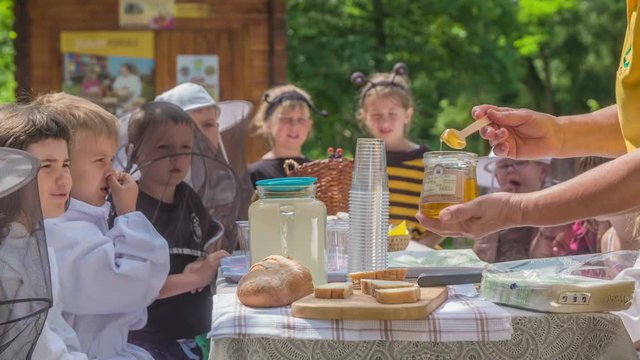 A teacher is giving honey on bread so that children will eat it. It's a really nice summer day and they are eating outdoors and are enjoying their snack.