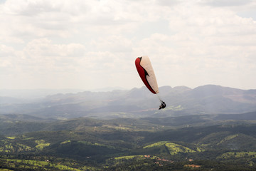 Lone paraglider gliding through the air over a green, mountainous backdrop and partially clouded skies.