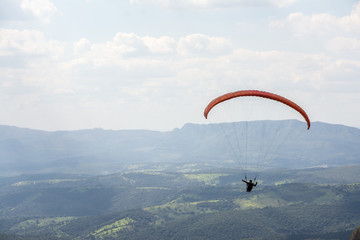 Lone paraglider gliding through the air over a green, mountainous backdrop and partially clouded skies.