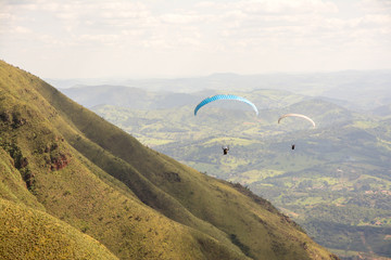Two paragliders gliding through the air over a green, mountainous backdrop and partially clouded...