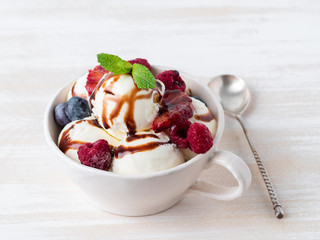 Balls of vanilla ice cream in mug with raspberry and blueberries, chocolate syrup  topping. Side view summer dessert, white, bright background