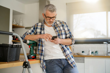 Mature man checking phone whilst fixing shelf in modern kitchen