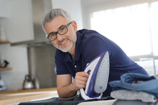 Mature man leaning on ironing board holding iron in modern kitchen