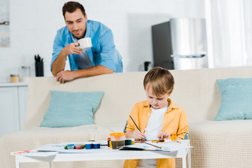 preschooler son sitting on couch and drawing at home while father drinking coffee on background