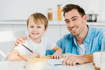 father and preschooler son looking at camera and drawing during breakfast in kitchen