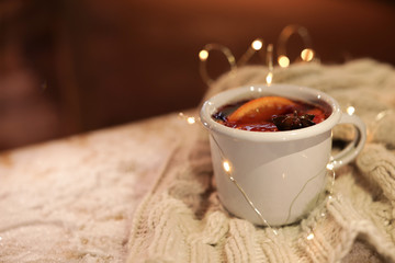 Cup of hot mulled wine and garland on table against blurred background. Space for text