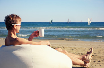 Male surfer in sunglasses resting on pouf on beach with cup of tea, in the background windsurfers...