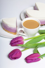 Fototapeta na wymiar Blueberry cheesecake. A piece of ready-made dessert on a saucer. Jelly layers of different colors are visible. Near a cup of coffee and a bouquet of tulips.